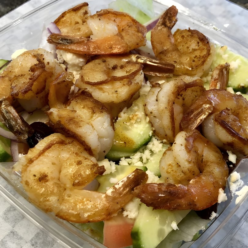 Close-up of a shrimp salad with cucumbers, feta cheese, and red onions in a plastic container.